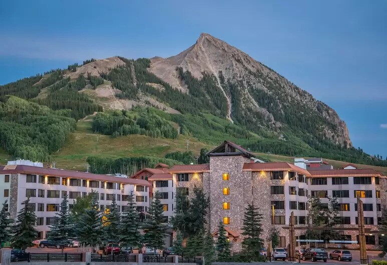 The Grand Lodge is depicted with its welcoming façade, five stories tall, situated against the backdrop of a majestic mountain. Surrounding trees add greenery to the scene, while warm lights glow softly from the building's windows at dusk.