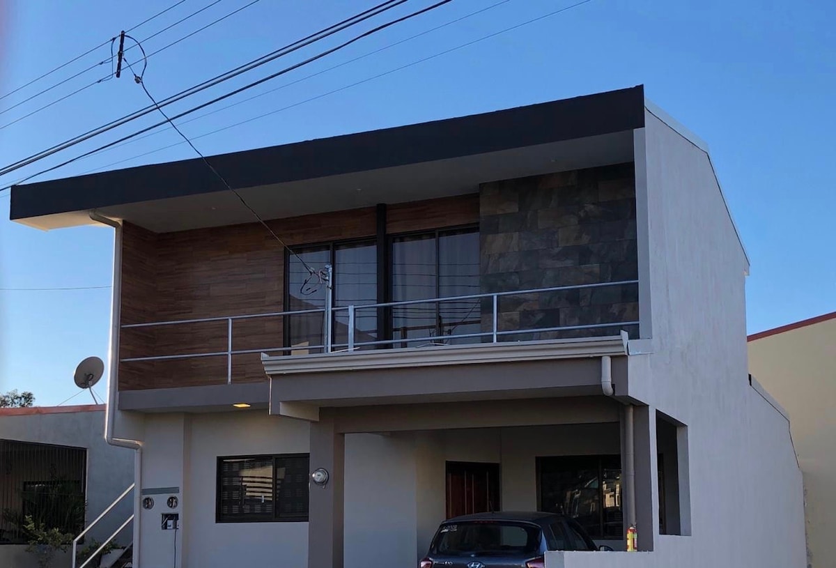 The exterior of the apartment is depicted, featuring a modern two-story design with a combination of wood and stone accents. Large windows allow natural light to enter, and a balcony extends from the upper floor. A parked car is visible in front of the building.