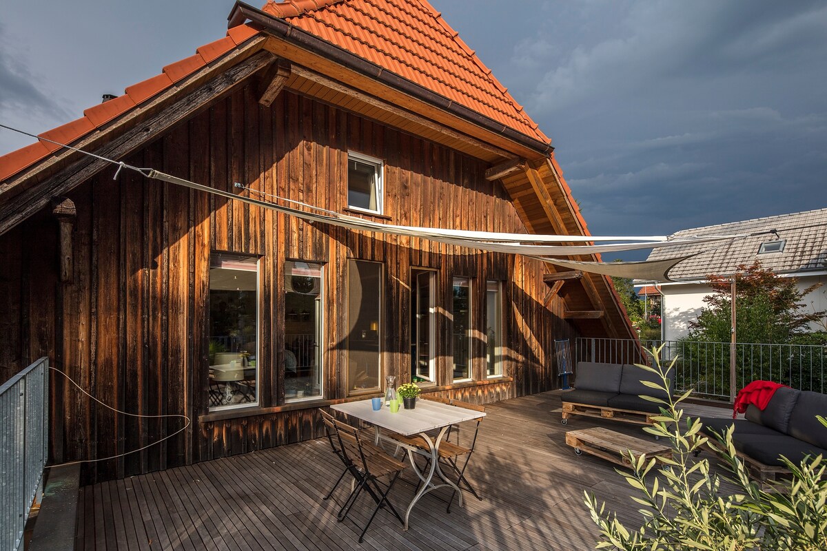 A large terrace features a wooden deck, complemented by a table and chairs. Two sofas are positioned nearby, inviting relaxation. The exterior showcases a traditional wooden facade with a red tiled roof, and large windows allow for ample natural light. Greenery adds a touch of nature.