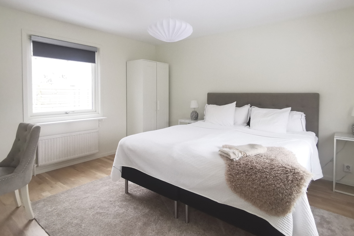 A spacious bedroom features a large bed dressed in white linens, complemented by soft pillows. Flanking the bed are two nightstands with lamps. A window allows natural light, and a cozy chair is situated nearby. A plush rug enhances the comfort of the room.