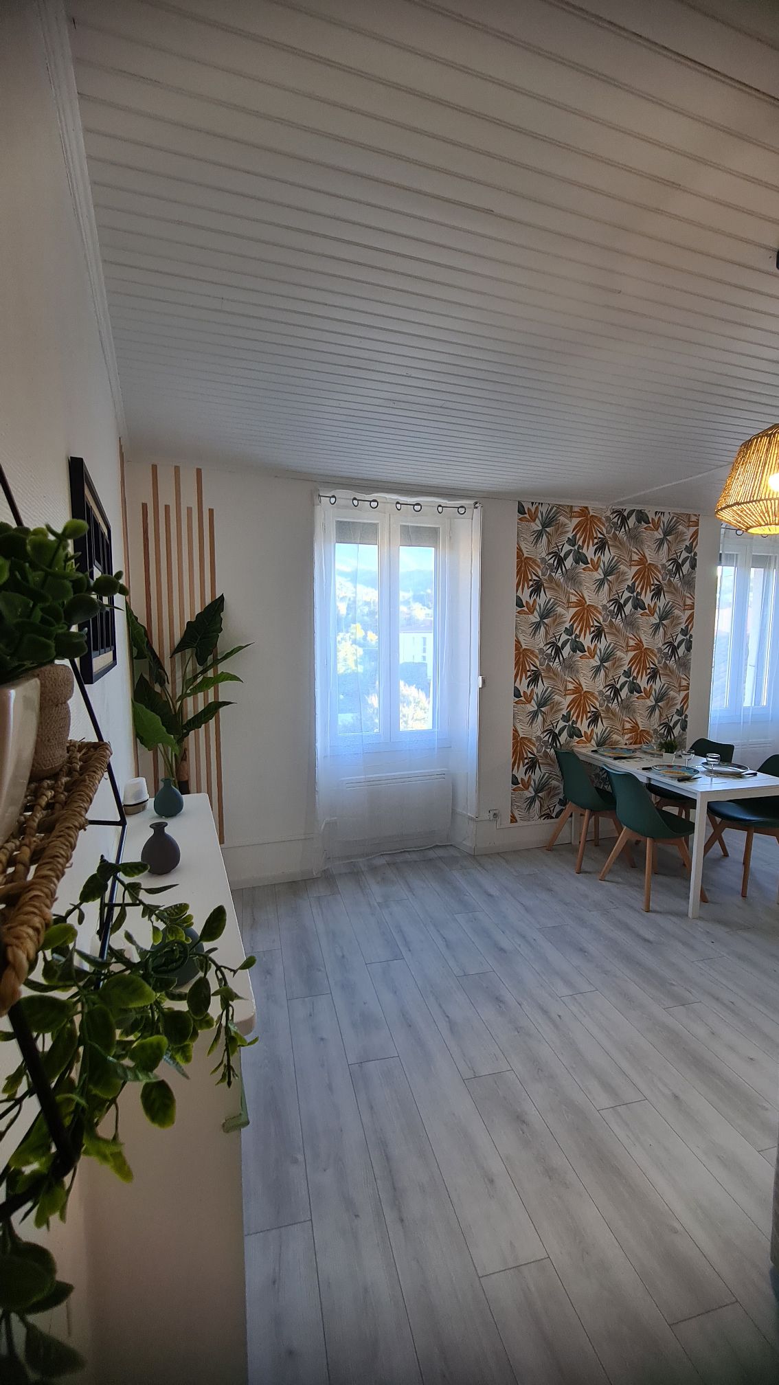 A bright and airy dining area features a large window adorned with light curtains, allowing natural light to flow in. A decorative wall with floral patterns complements a clean, modern dining table surrounded by teal chairs. Greenery from potted plants adds a touch of color.