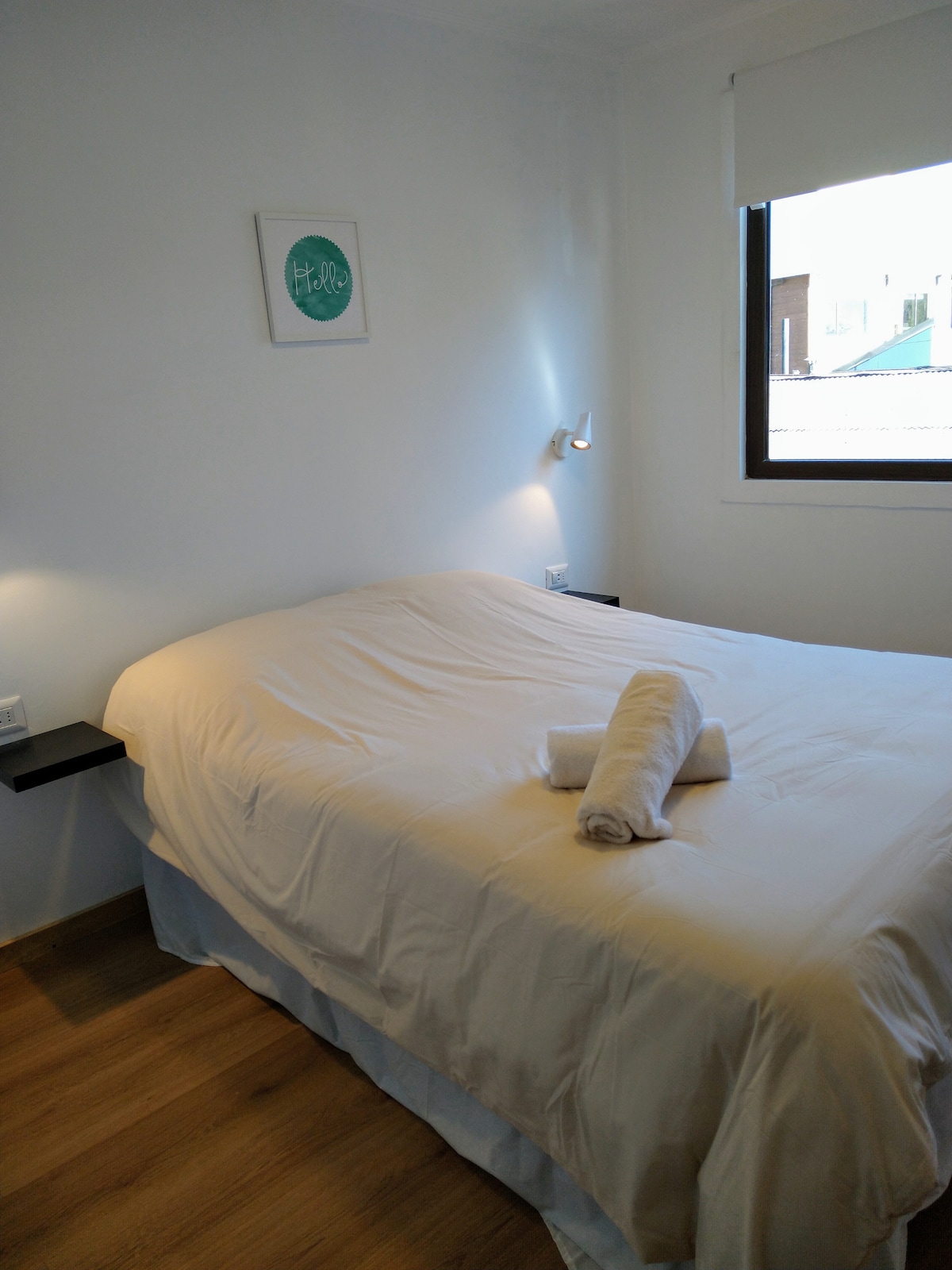 A light-filled sleeping area features a spacious bed with a neatly made white duvet. A rolled towel is placed on the bed for convenience. The room is complemented by modern wall sconces and a large window with a minimalistic blind.