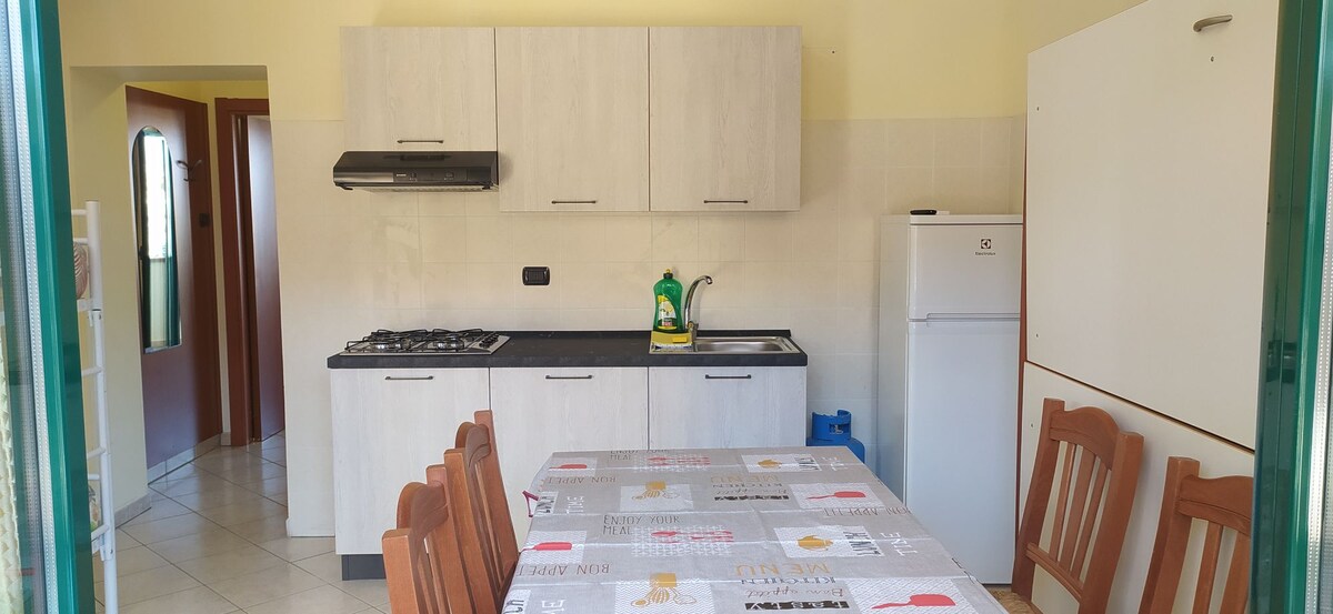 A clean and functional kitchen area is presented, featuring a wall-mounted range hood above a gas stove. Counter space is available with cleaning supplies and a sink. A dining table, surrounded by wooden chairs, is positioned in the center, enhancing the communal atmosphere.