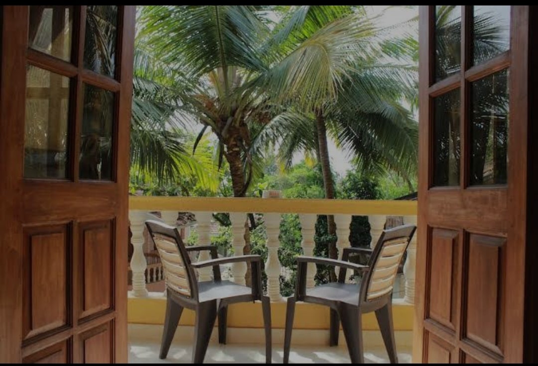 A balcony view is presented, featuring two comfortable chairs positioned on a smooth surface. Lush palm trees and greenery are visible in the background, allowing natural light to filter through the open wooden doors.