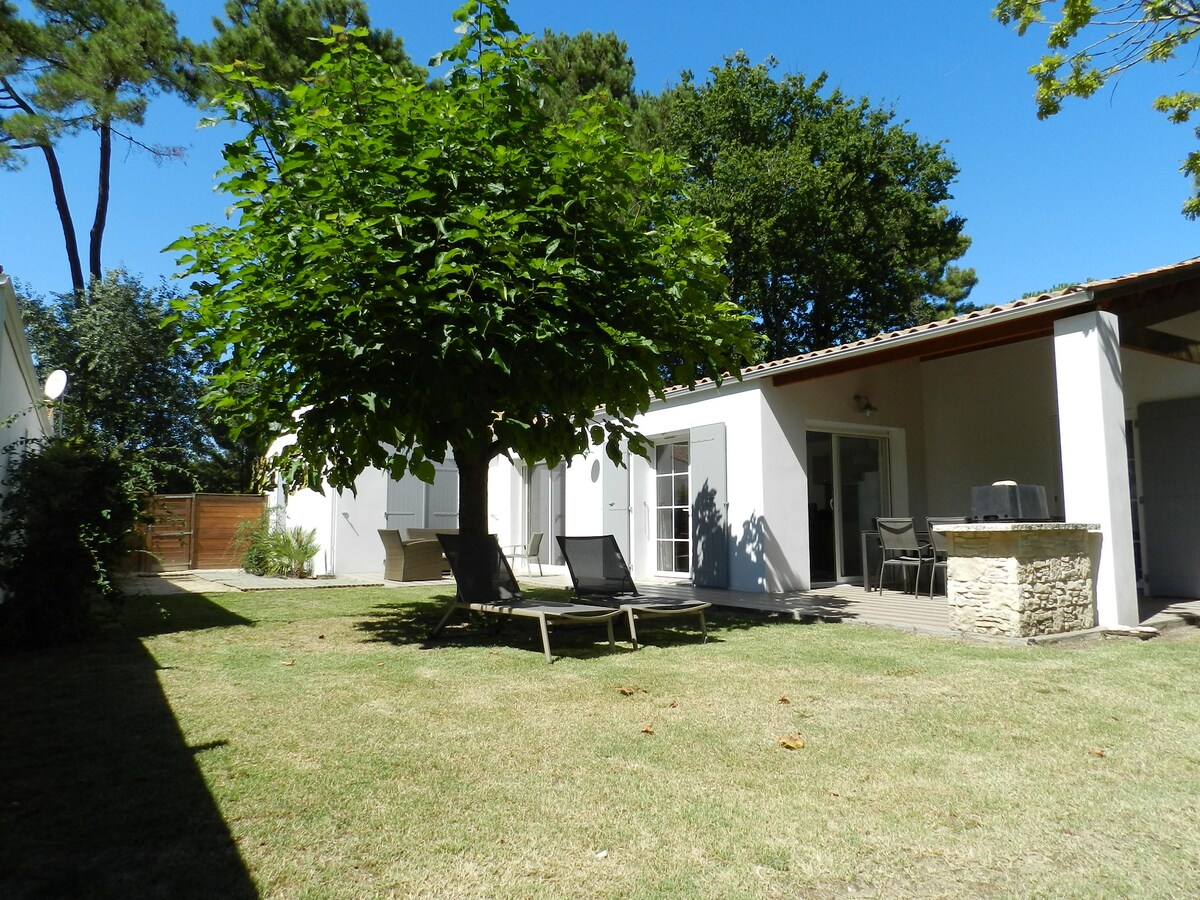 A private garden area features a shaded lounge space with sun loungers, surrounded by greenery. The villa's exterior is visible, showcasing large glass doors and a covered terrace with a barbecue setup. The flat lawn provides ample space for relaxation.