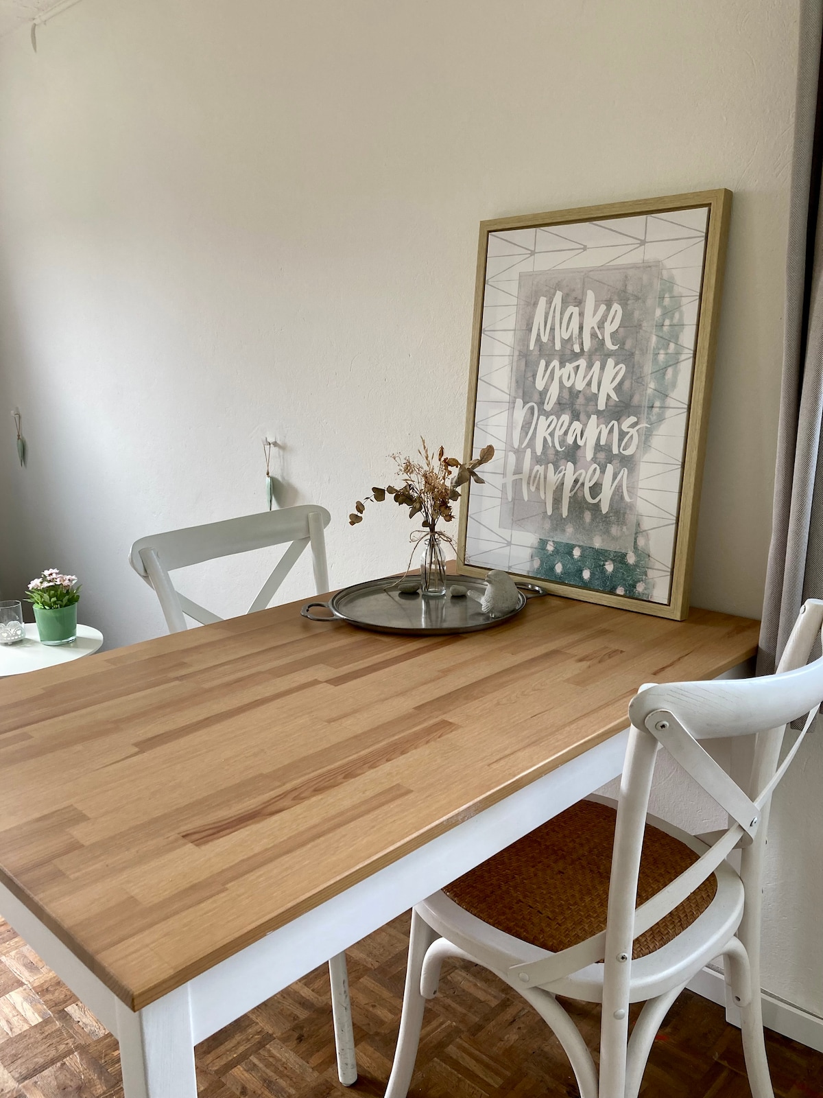 A light wooden table is complemented by two white chairs, enhancing the dining space. A decorative framed poster is located on the wall, with the text 'Make your Dreams Happen.' A small plant and dried flowers are placed on the table, adding a natural touch.