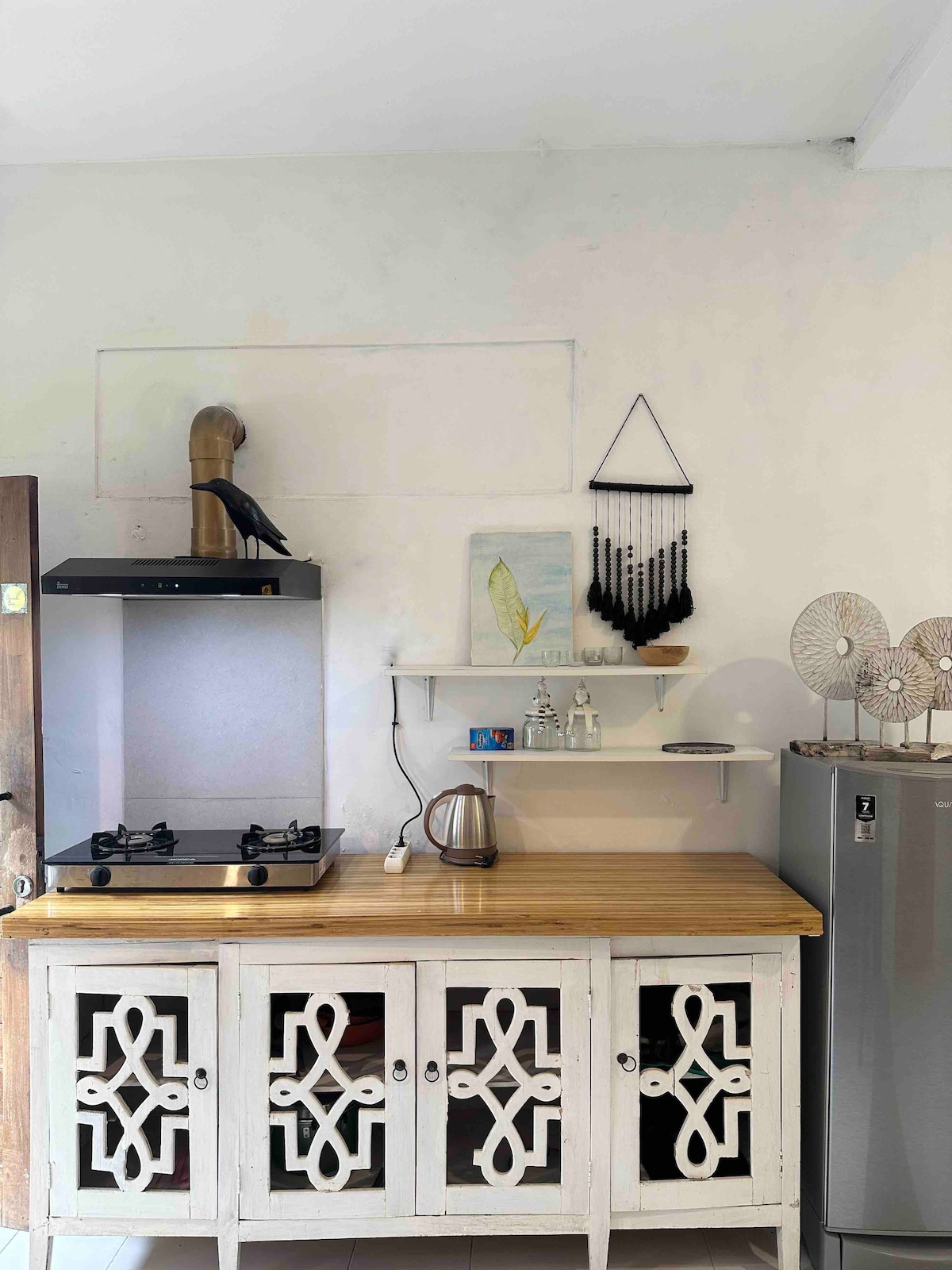 A functional kitchen space is depicted, featuring a wooden countertop and a modern stovetop. The minimal decor includes a kettle, small art piece, and handcrafted wall hangings. A sleek refrigerator stands nearby, emphasizing the clean and simple design of the area.