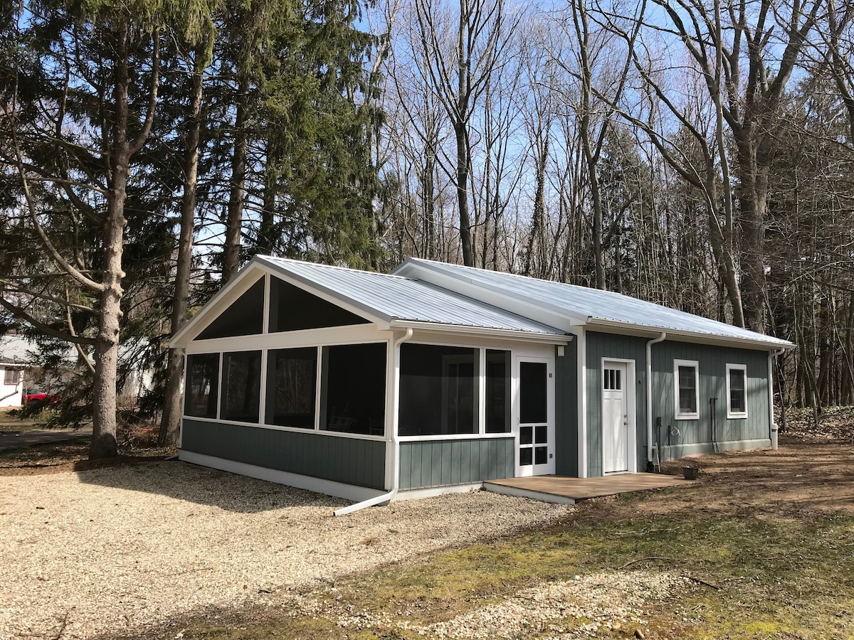 The exterior of a modern guest house is visible, featuring a combination of green siding and a metal roof. Large screened-in porch sections offer ample outdoor space, surrounded by trees and gravel landscaping. A wooden deck extends from the side entrance.