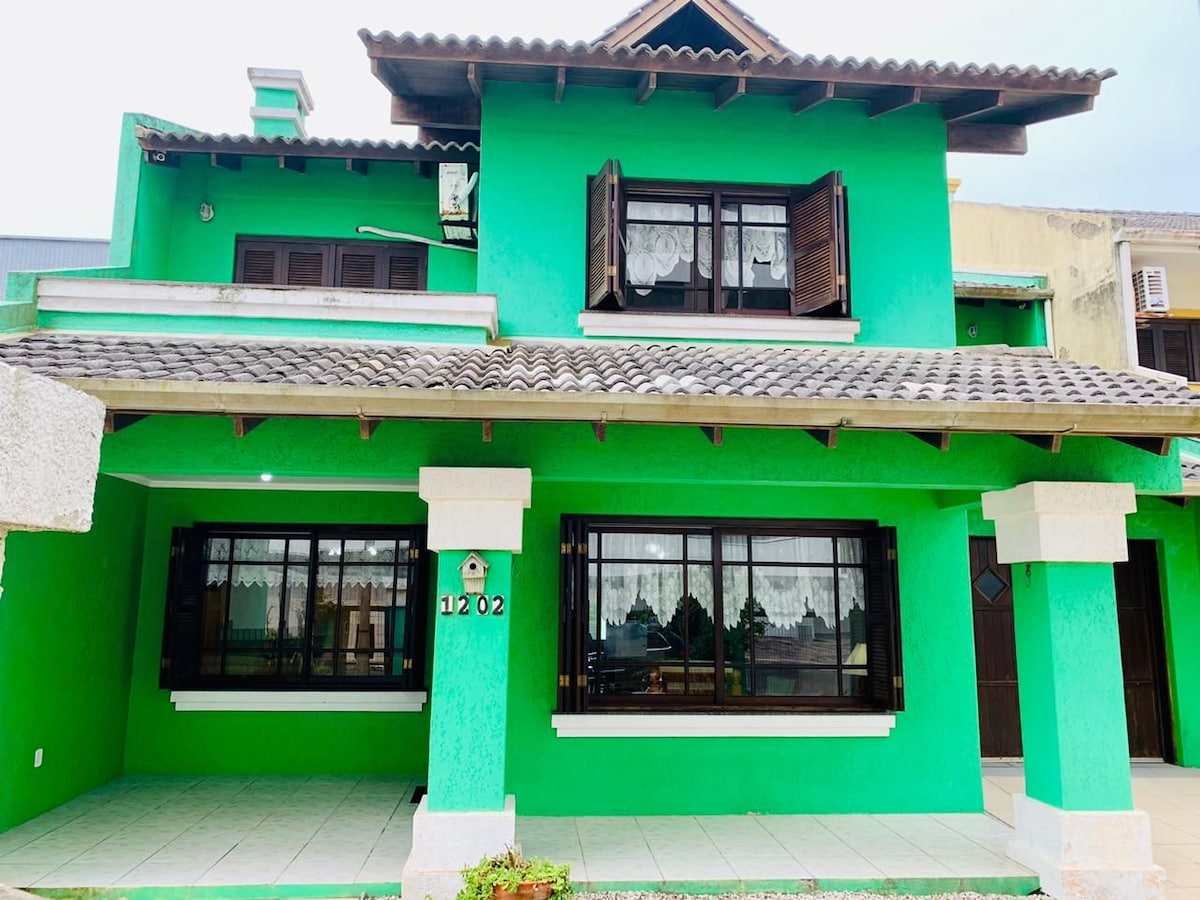 The exterior view showcases a two-story house painted in vibrant green. Characterized by wooden windows and a tiled roof, it features a covered entrance with a stone façade. The surrounding area is paved, complemented by neatly maintained vegetation adding to the inviting appearance.