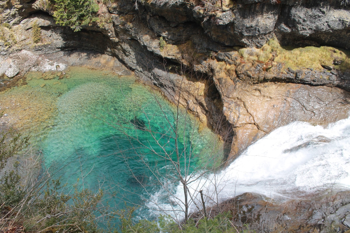 A serene natural pool is nestled among rocky cliffs, featuring clear turquoise waters gently flowing into a small waterfall. The surrounding area is adorned with patches of greenery, creating a tranquil and inviting landscape.