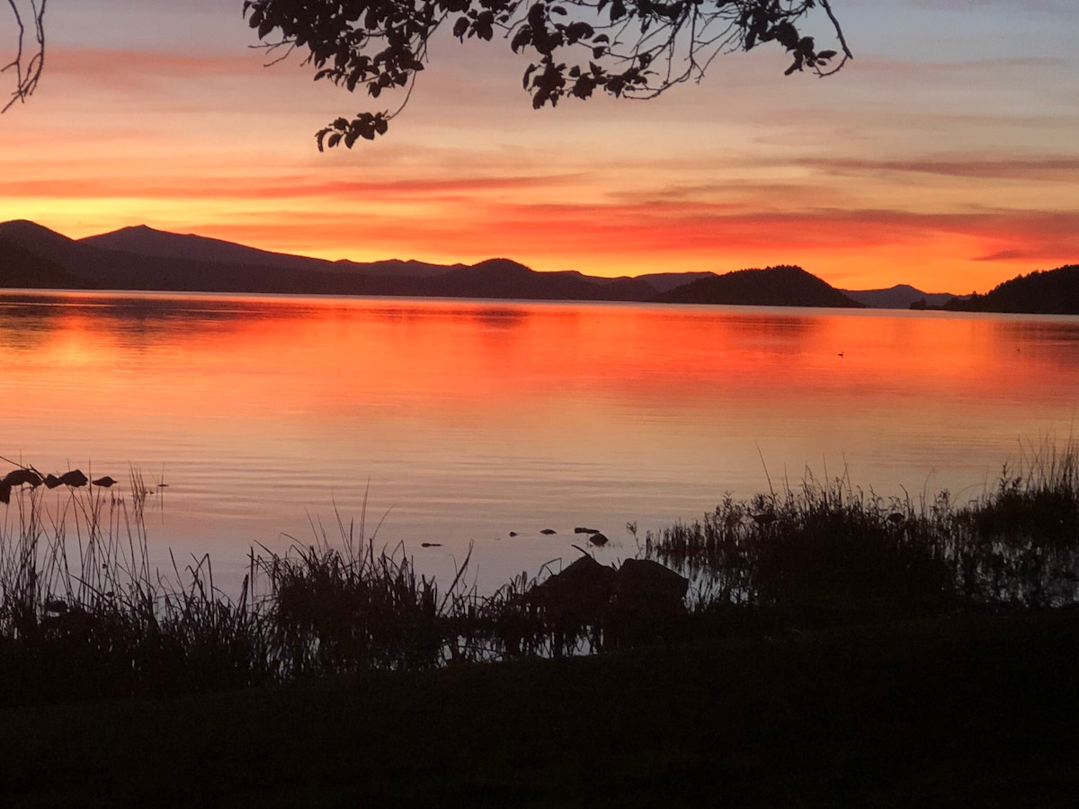 A tranquil sunset over Klamath Lake showcases vibrant hues of orange and pink reflecting on the water's surface. Silhouetted mountains are visible in the background, with gentle grasses lining the foreground, creating a serene lakeside scene.