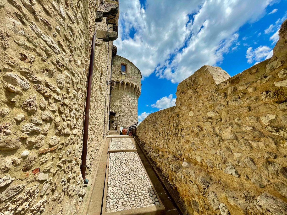 An outdoor pathway is seen, lined with smooth stone and framed by ancient stone walls. The pathway leads toward a round tower, bathed in natural light under a blue sky dotted with clouds. The scene conveys a sense of history and tranquility.