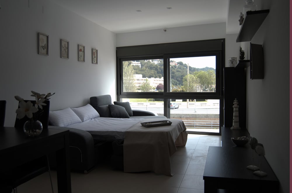 A modern living area is displayed, featuring a spacious sofa bed and a chaise longue. Large windows allow natural light and offer views of the surrounding greenery. Elegant wall art and minimalist furniture complement the uncluttered aesthetic.