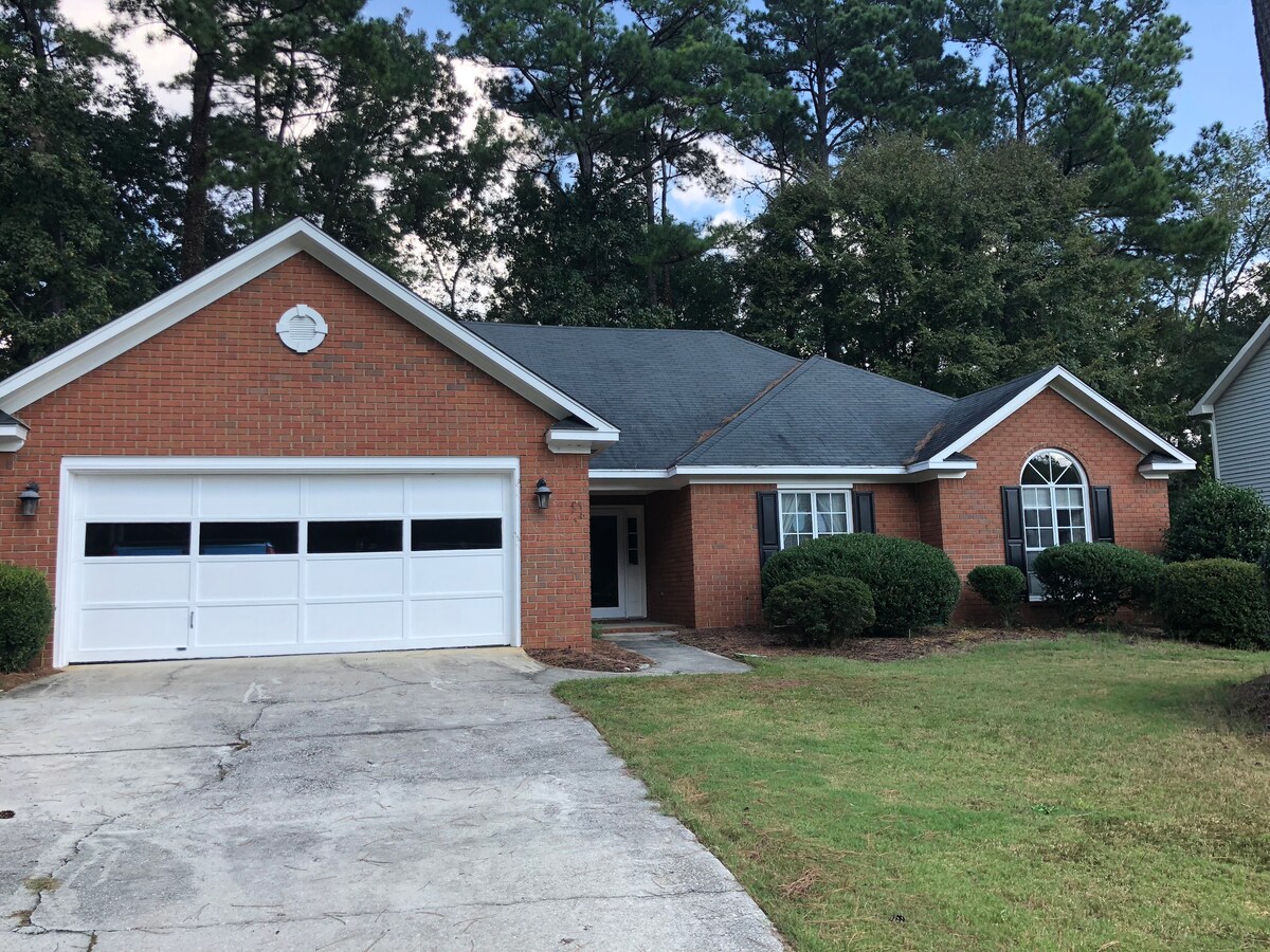 A single-story brick home is showcased, featuring a two-car garage with a white door. Lush hedges frame the front yard, and a concrete driveway leads to the entrance. Large windows flank the front door, inviting natural light into the interior.