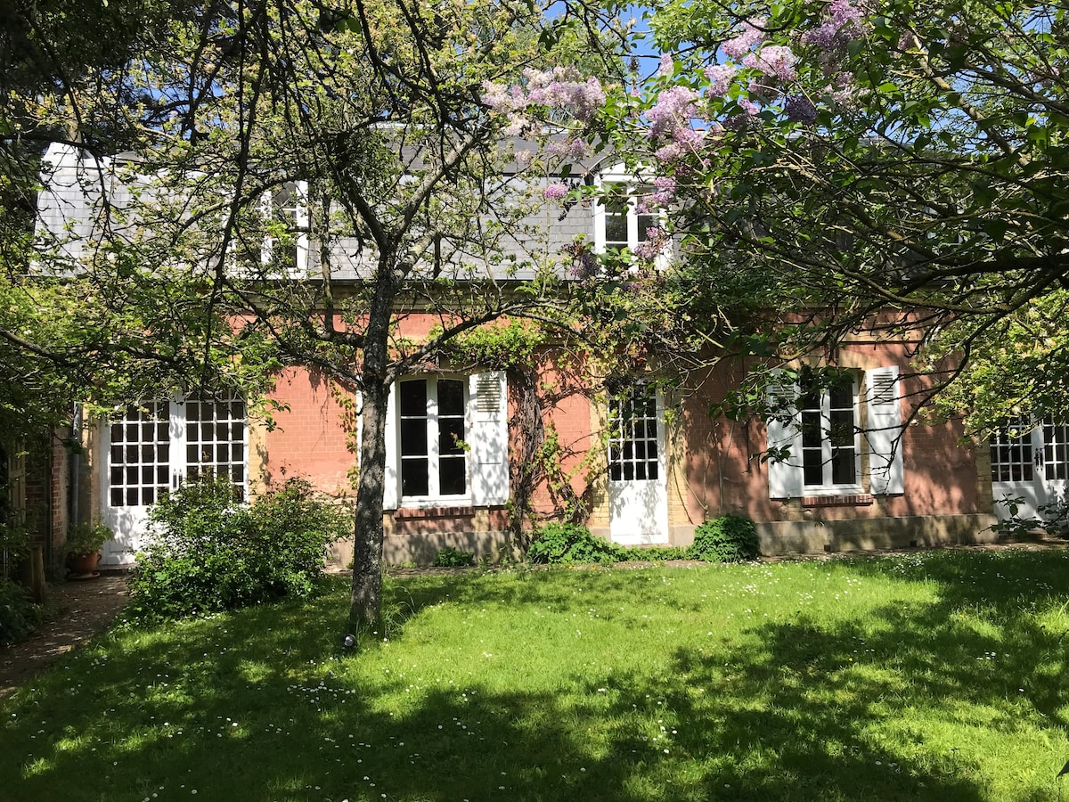 A charming house is nestled within a lush garden, featuring mature trees and flowering plants. Large windows reflect natural light, offering views of the serene outdoor space. The well-maintained lawn is framed by vibrant greenery, creating a tranquil environment.