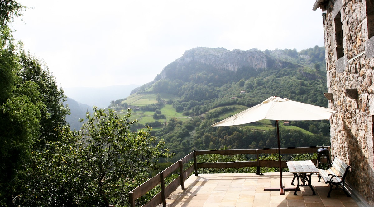 A spacious terrace features a large umbrella providing shade over a picnic table. Lush green hills and mountains stretch into the distance, offering a peaceful natural landscape. Stone walls of the house add rustic charm to the outdoor area.