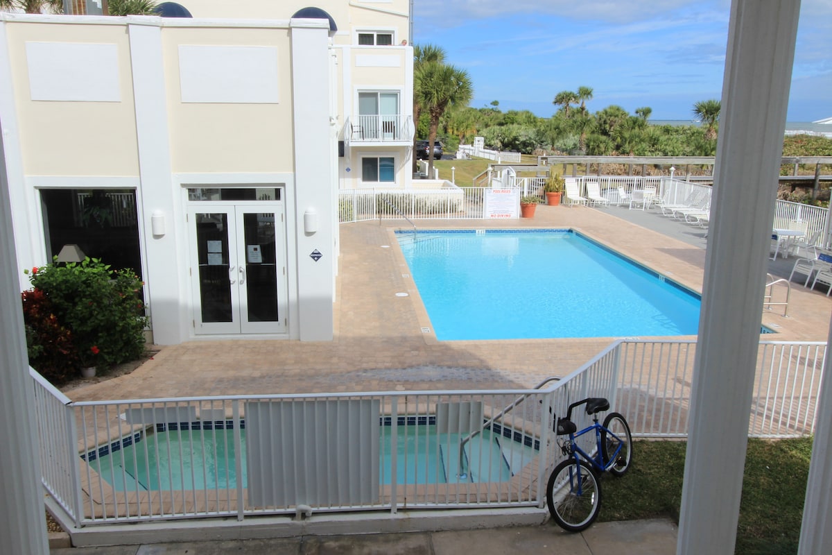 A view of the community pool area is presented, featuring a large swimming pool alongside a smaller hot tub. Sun loungers are arranged around the pool, with palm trees visible in the background, and the beach is visible beyond the pool area.