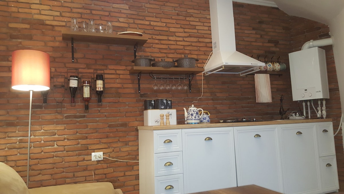A kitchen area features exposed brick walls, enhancing the rustic feel. The countertop is equipped with cooking essentials, including a stove and varied cookware. Above, glass shelves display glassware and a decorative tea set. A warm lamp provides soft lighting, completing the cozy atmosphere.