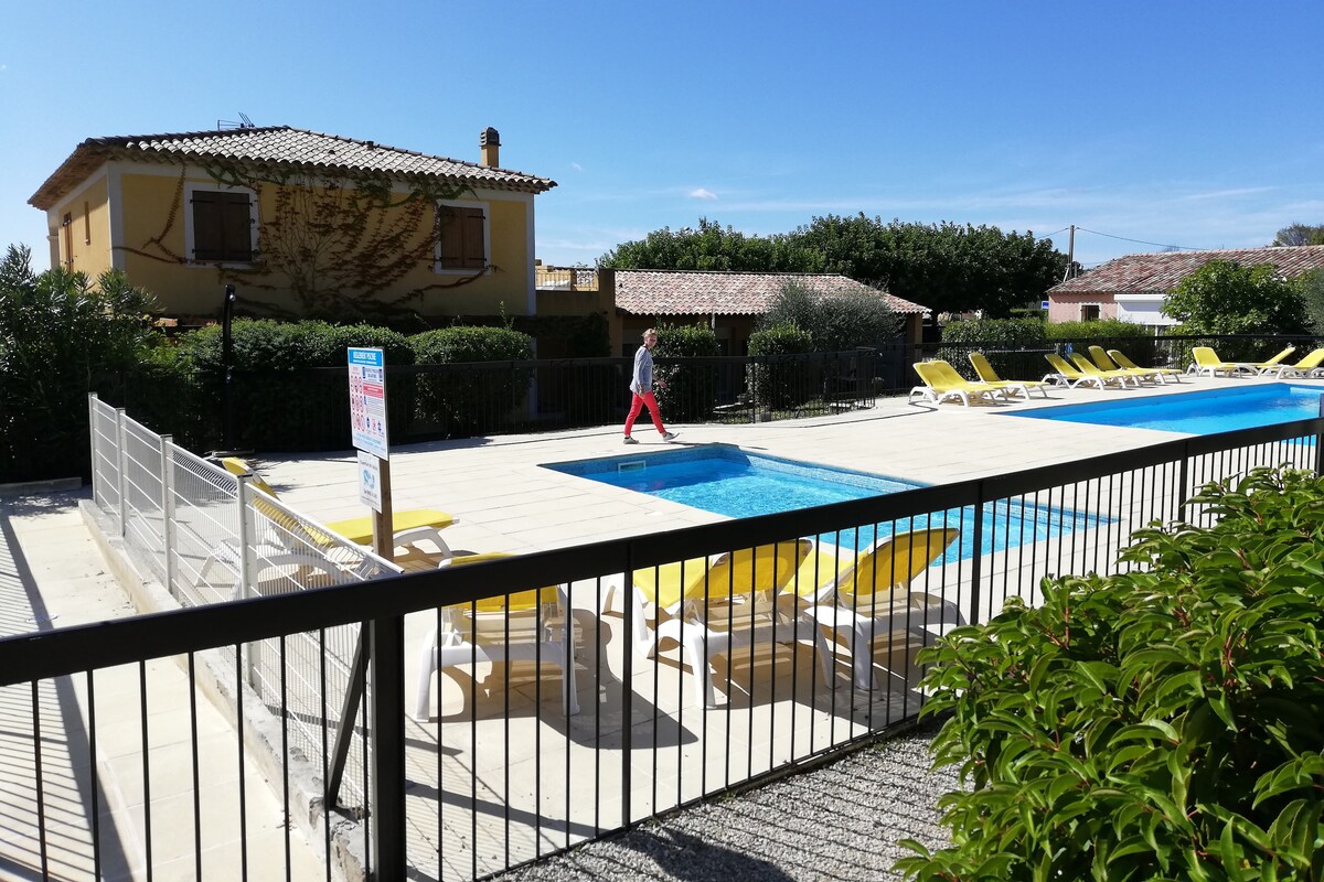 An inviting pool area is surrounded by sun loungers and greenery. The clear water reflects the blue sky above, while a person walks along the edge of the pool. A yellow house with a terracotta roof is visible in the background.