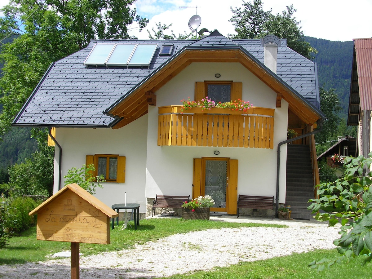 The exterior of La Casetta is depicted, featuring a two-story structure with orange window frames and a wooden balcony adorned with flowers. A welcoming garden area with greenery surrounds the entrance, while a wooden sign marks the property's name. The backdrop showcases the serene mountain landscape.