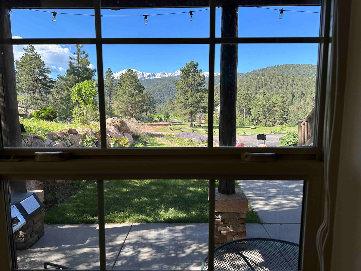 A view from a window reveals a scenic landscape featuring a garden area, rocky outcrops, and trees. Mountains are visible in the background under a clear blue sky. Outdoor seating is present on the patio, offering a peaceful atmosphere.