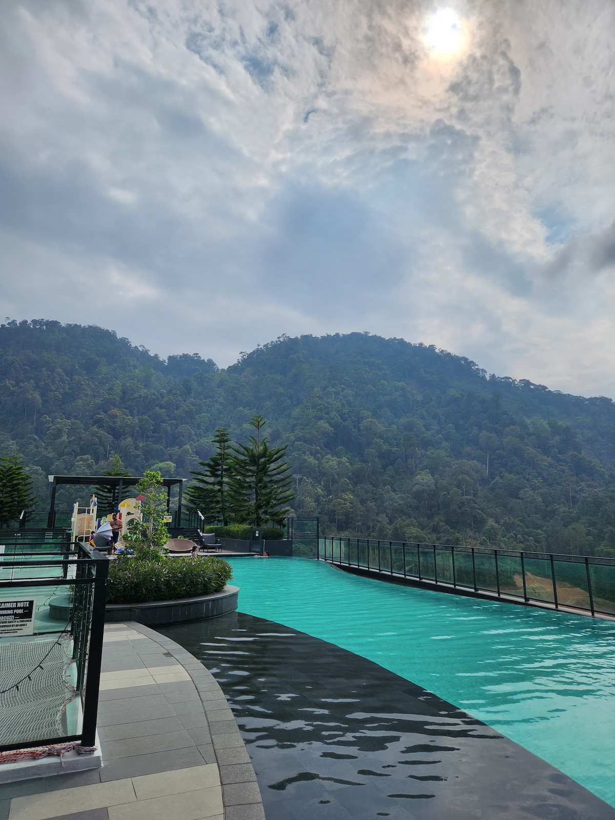 The image showcases an expansive swimming pool with a tranquil turquoise hue, surrounded by verdant mountains under a cloudy sky. The pool area features a sleek design with a gentle curve, and lounge seating is visible in the background, inviting relaxation amidst the natural scenery.