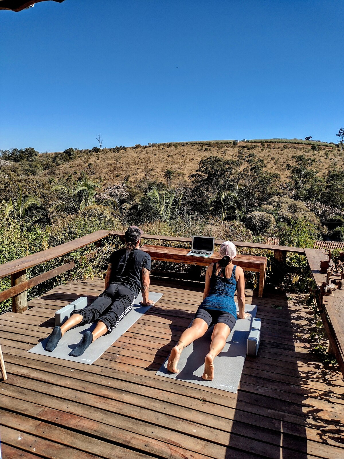 Two individuals are engaging in yoga on a wooden deck, overlooking a scenic landscape filled with greenery and rolling hills. A laptop is positioned nearby, and clear blue skies are visible above, enhancing the tranquil outdoor setting.