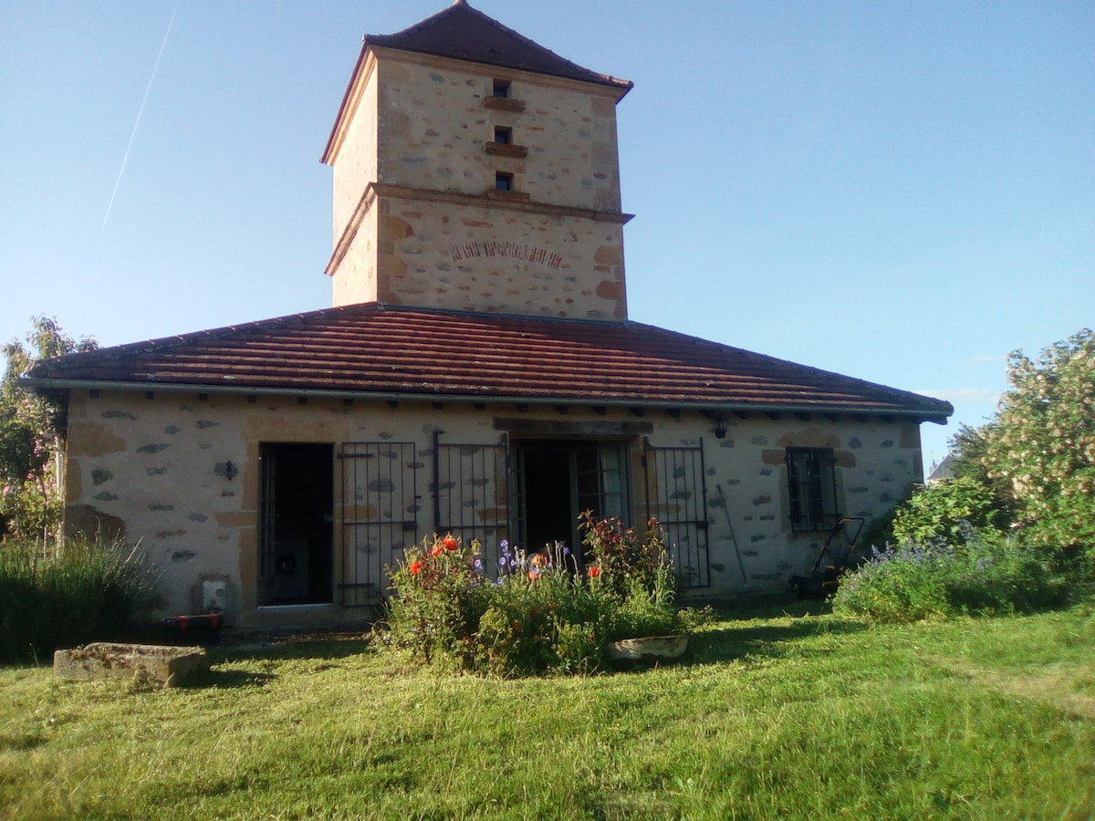 The exterior of a renovated pigeonnier is visible, characterized by natural stone walls and a traditional roof. The surrounding grassy area is adorned with a variety of flowering plants, and large windows frame the entrance, complementing the tranquil rural setting.