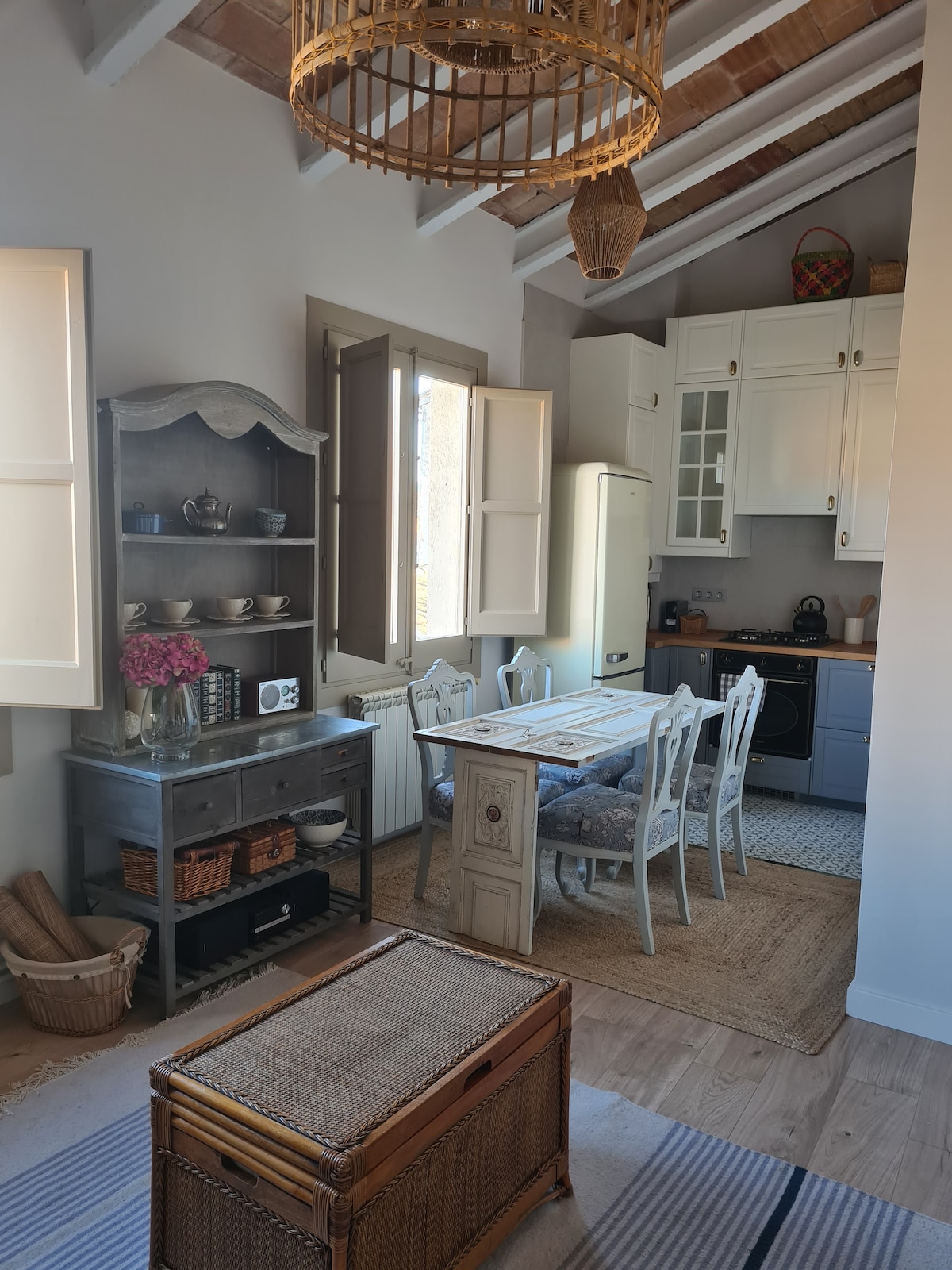 A warm, inviting kitchen area features a wooden dining table surrounded by six chairs. A vintage cabinet holds dishware, and fresh flowers add a touch of color. The space is illuminated by natural light streaming through open shutters, highlighting the charming wooden ceiling.
