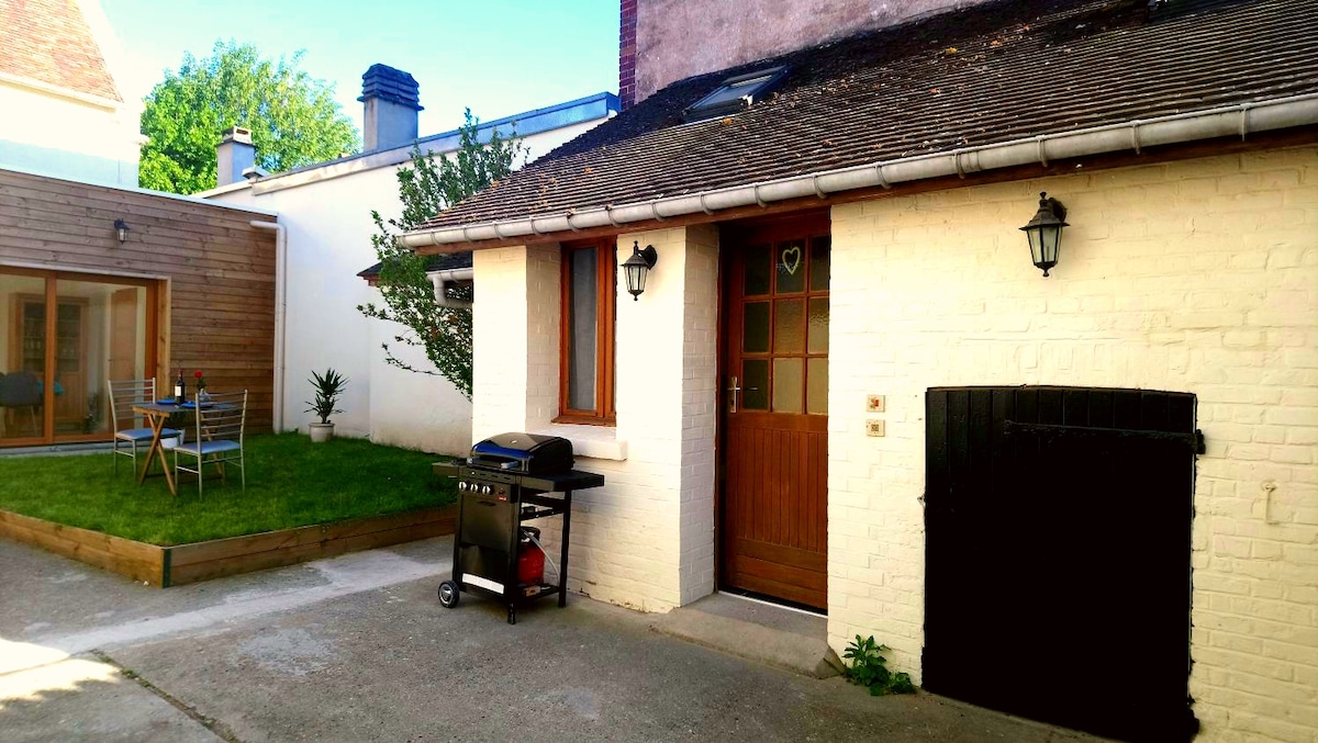 A well-maintained outdoor space is presented, featuring a small garden area with a wooden platform and a set of patio chairs. A charcoal grill is positioned near the entrance, with a traditional door offering access to the dwelling. Natural light illuminates the space, surrounded by greenery.