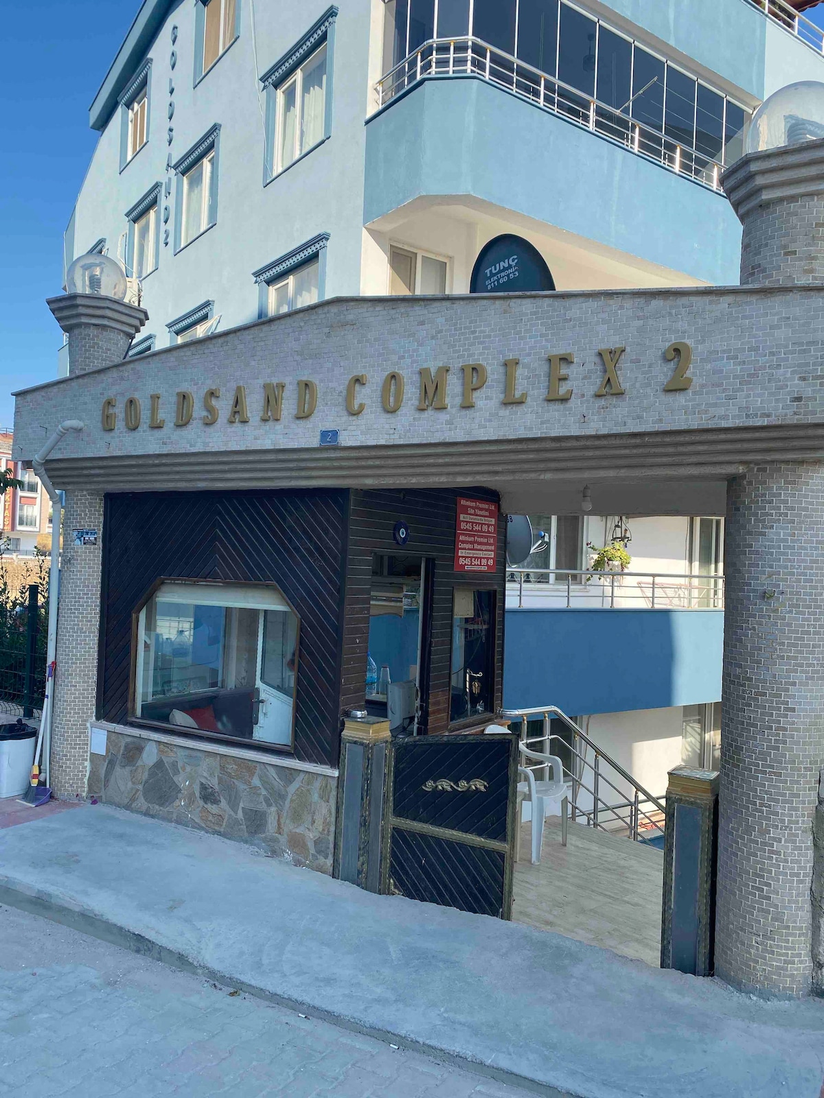 The entrance to Goldsand Complex 2 features a stylish facade with the name prominently displayed. A stone front and wooden accents complement the modern design. The surrounding area includes outdoor seating and a glimpse of the building’s upper levels beyond the entrance.