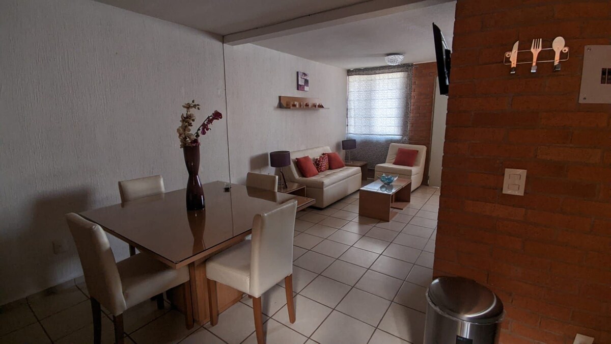 A bright living area features a simple dining table surrounded by four chairs, with a comfortable sofa adorned with colorful cushions. Natural light floods in through a window, illuminating the tiled floor and light-colored walls.
