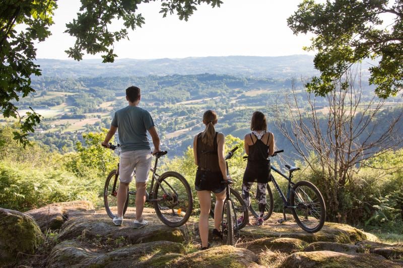 Three individuals stand on a rocky overlook, gazing at a vast landscape of rolling hills and valleys. Mounting mountain bikes, they are framed by trees and greenery, suggesting a serene and natural environment. Soft light enhances the tranquil scene.