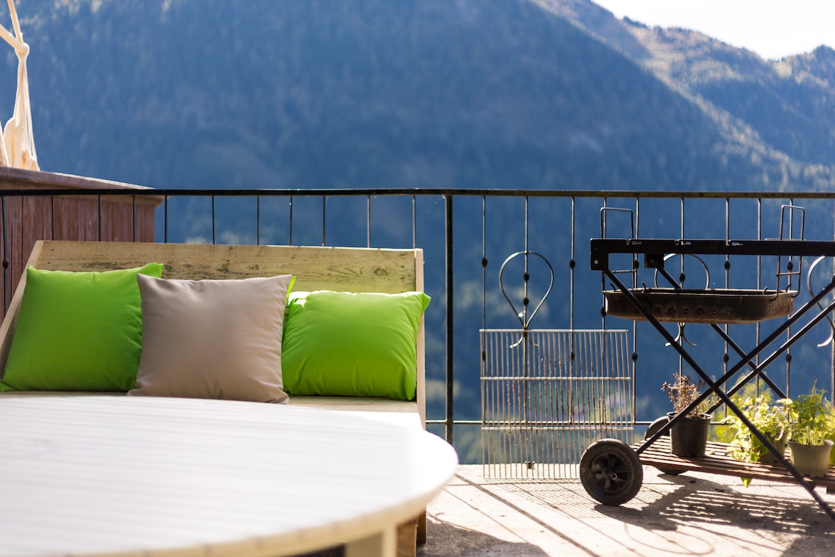 A comfortable outdoor seating area features a circular table surrounded by pillows in natural tones, with green accents. A charcoal grill is positioned nearby, offering a view of the mountains through a balcony railing. The serene landscape enhances the peaceful atmosphere.