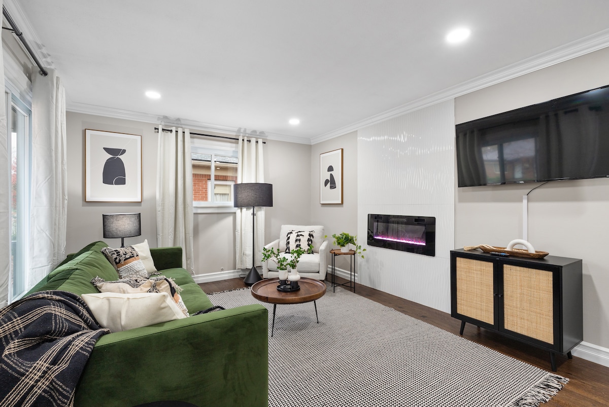 A cozy living area features a green sofa paired with decorative pillows. A round table, adorned with a plant, sits in front of the sofa. A wall-mounted TV and an electric fireplace add warmth, while natural light filters through the window covered by light curtains.