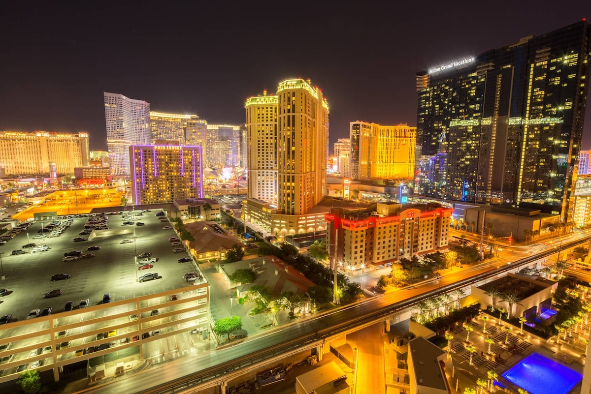 An aerial view captures the vibrant Las Vegas Strip illuminated at night, showcasing an array of hotels and casinos. The scene includes a parking facility and the scenic monorail track, with sparkling lights creating a lively atmosphere throughout the cityscape.