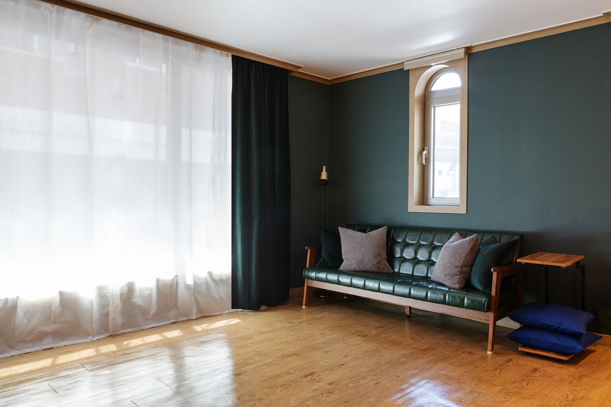 A living area features a green leather sofa adorned with two decorative pillows. Soft natural light streams through a large sheer curtain beside a smaller window. A wooden side table sits beside the sofa, offering a quiet, functional space against a muted dark green wall.