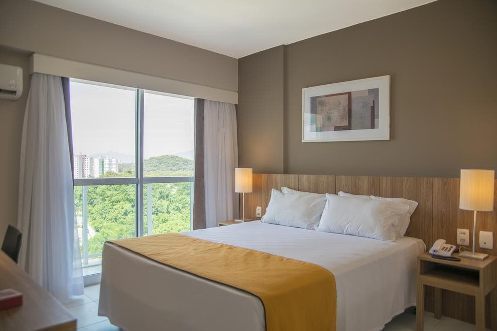 The room features a spacious bed with crisp white linens and soft pillows, complemented by a mustard-colored throw. Two bedside lamps provide gentle lighting, and a large window offers views of the surrounding greenery. A light wooden headboard and desk enhance the modern design.