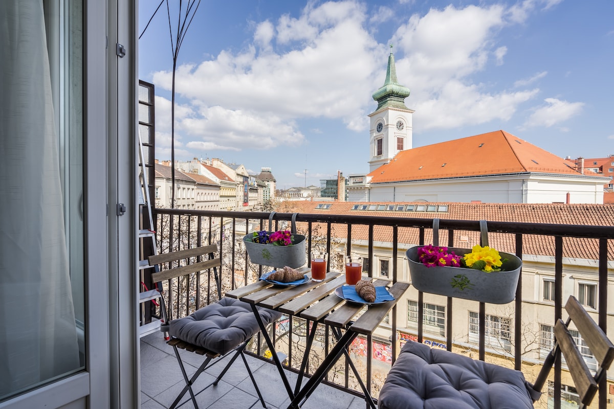 A small balcony features a table set for two, adorned with colorful flower pots and breakfast items. A view of historic buildings and a church tower is visible against a clear blue sky, providing a charming backdrop for outdoor dining.