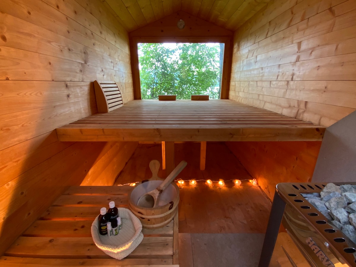 A wooden sauna interior features warm lighting along the floor and two benches positioned near a large window, offering a view of greenery. The space includes a basket with essential oils and a wooden spoon, adding relaxation elements to the serene environment.