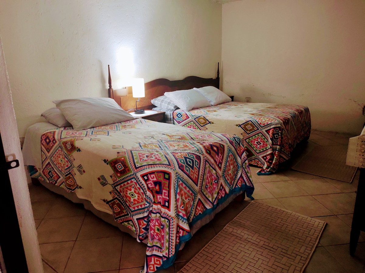 The room features two spacious beds, each covered with colorful patterned bedspreads. A bedside table with a lamp is positioned between the beds, providing soft lighting. The floor is tiled and a small area rug is present, contributing to the overall comfort of the space.