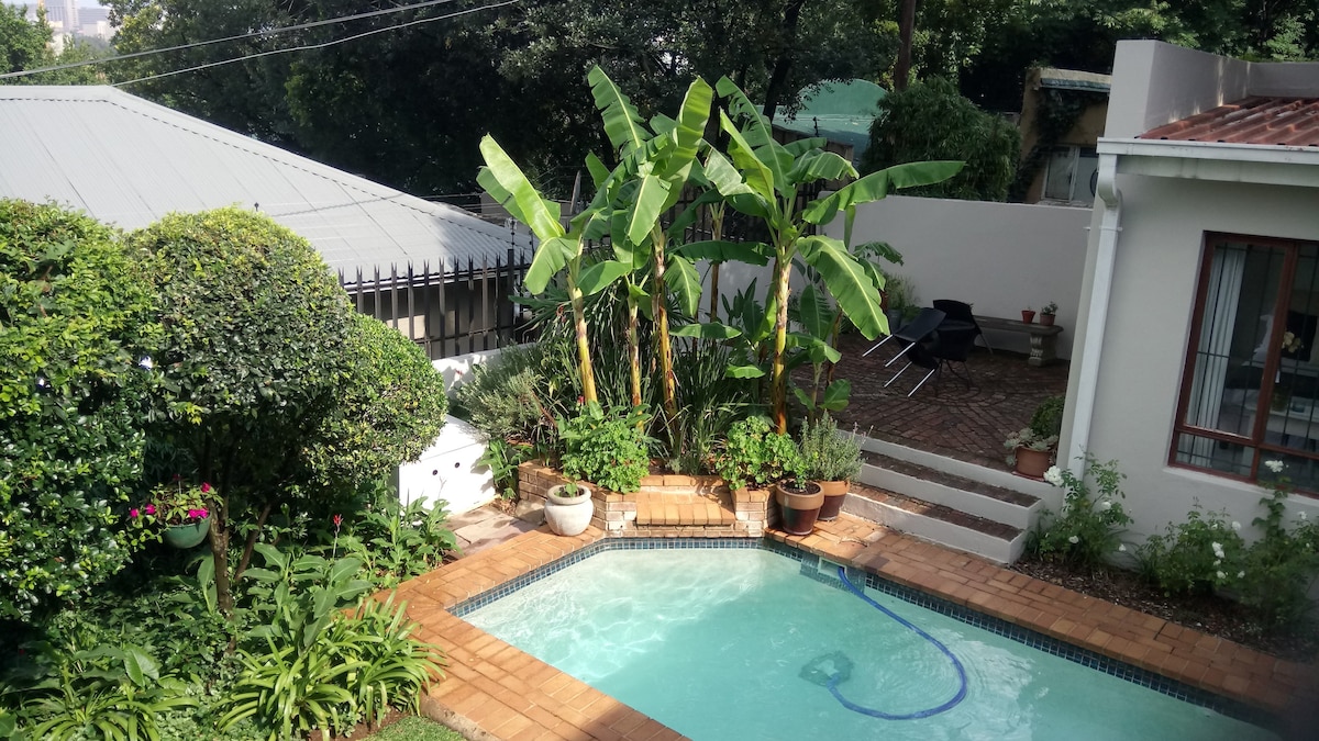 A private outdoor area features a swimming pool surrounded by lush greenery and potted plants. Tall banana plants create a tropical feel, while a patio with a pathway leading to the cottage is visible in the background.