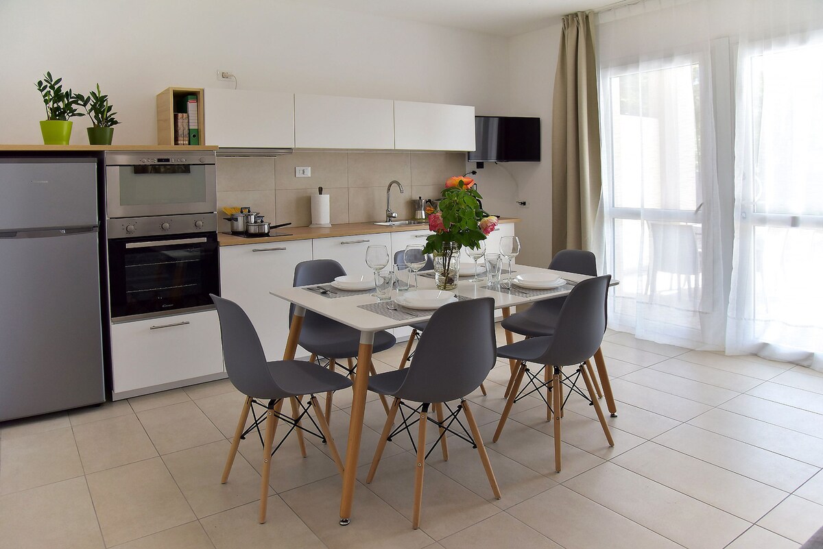 A modern kitchen area showcases a minimalist design featuring a dining table surrounded by six gray chairs. Kitchen appliances, including a refrigerator and oven, are visible along with a potted plant on the counter. Natural light filters through sheer curtains, enhancing the spacious feel.