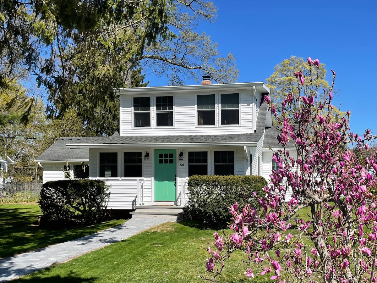 A well-maintained two-story house is framed by lush greenery, featuring a light-colored exterior and a distinctive mint green front door. Flowering shrubs are positioned in the foreground, adding a touch of color to the inviting entrance. Clear blue skies provide a bright backdrop.