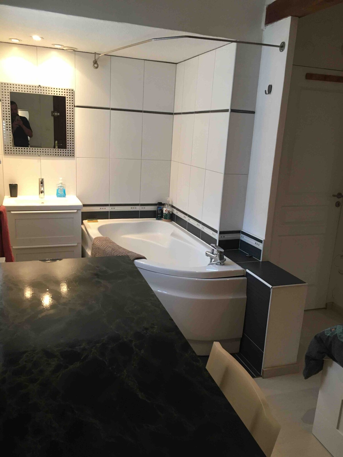 A spacious bathroom is presented, featuring a modern bathtub with a sleek design. A large mirror reflects ambient light, while a dark countertop contrasts with the white tiles. Storage options are available with cabinets, enhancing overall functionality.