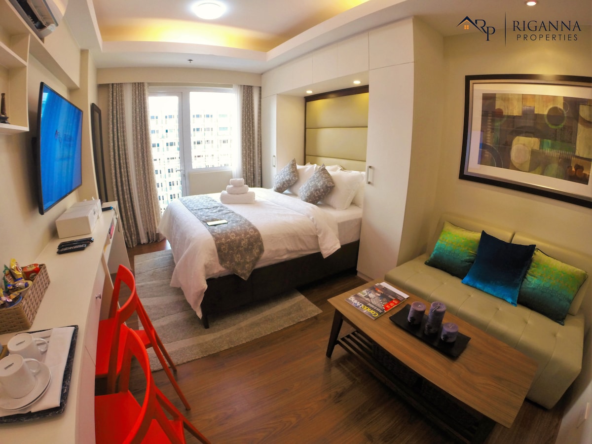 A well-appointed studio unit features a spacious bed with a decorative throw, complemented by a modern sofa in a neutral tone. A television is mounted on the wall, while a small dining area with vibrant red chairs is seen in the foreground. Natural light filters through the window, illuminating the room.