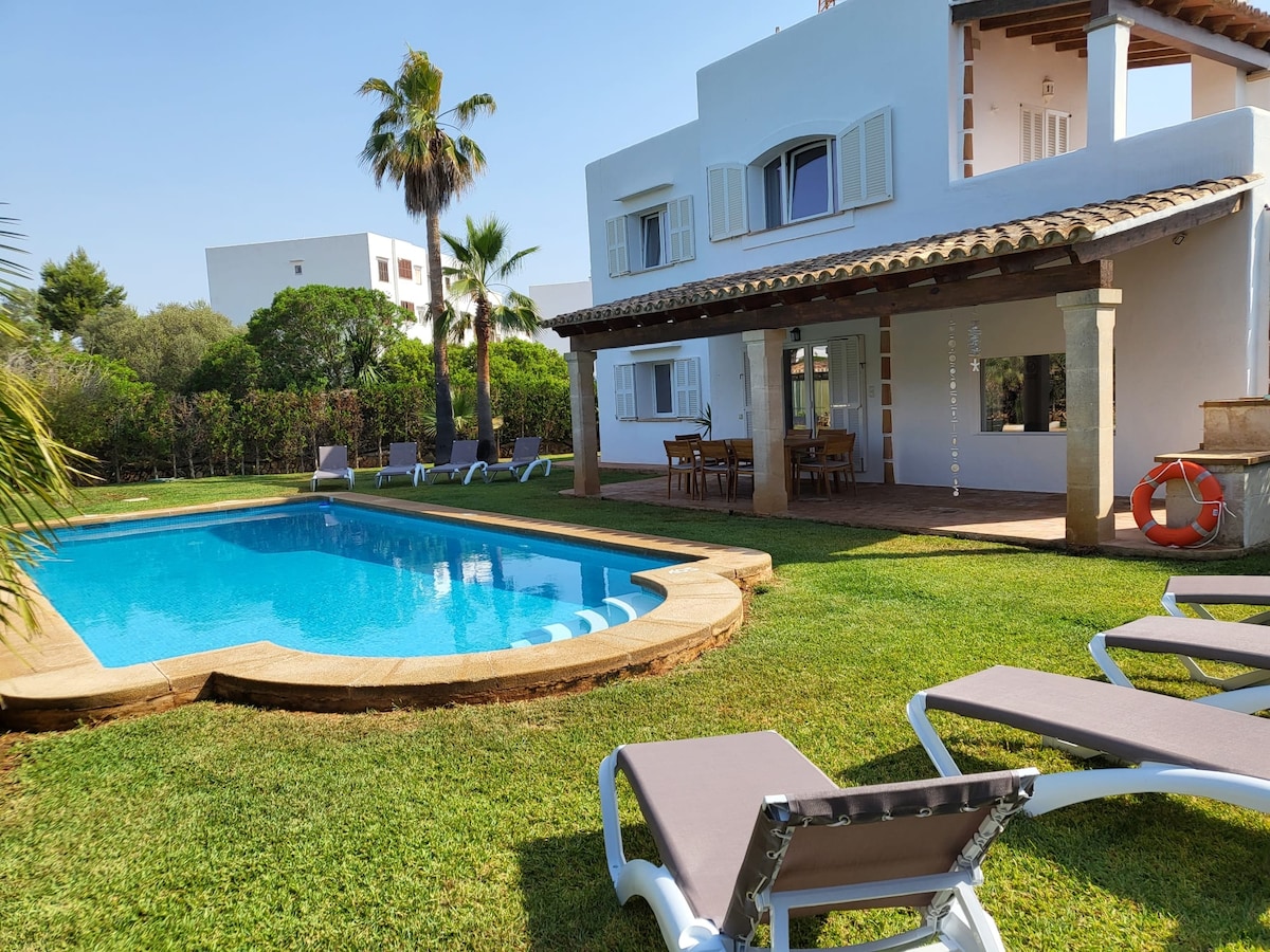 A private pool, measuring 10 by 5 meters, is surrounded by a well-maintained lawn and lounge chairs. A covered terrace features a dining table, and palm trees provide shade in the background. The two-story villa showcases charming architectural details and spacious outdoor areas.