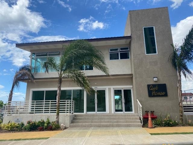 A modern clubhouse features large glass windows that allow natural light to fill the space. The entrance is flanked by palm trees and a pathway leading up to double doors. A sign reading 'Club House' is mounted on the building's exterior.