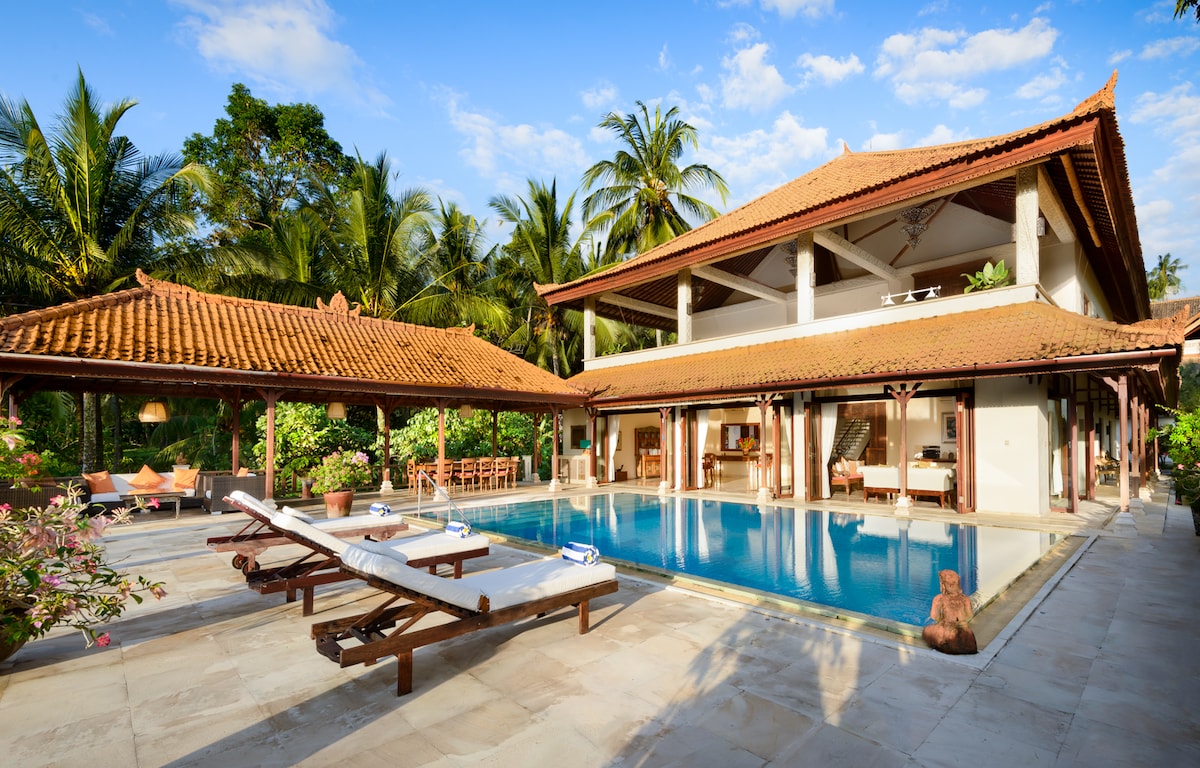 An expansive outdoor area features a tranquil swimming pool surrounded by lounge chairs. Lush tropical greenery provides a natural backdrop, while traditional Balinese architecture, characterized by its thatched roofs and open spaces, is prominently displayed. Multiple seating areas are available for relaxation.