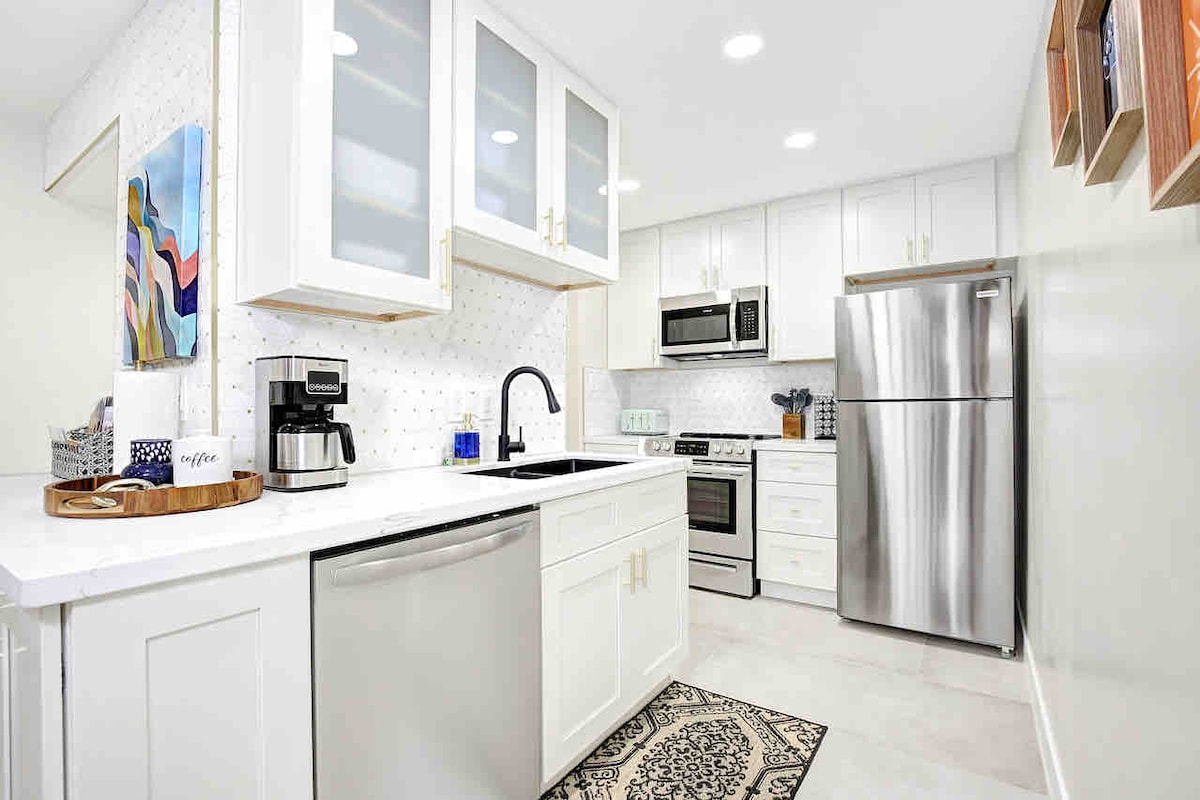 The modern kitchen features white cabinetry with glass accents, stainless steel appliances, and a decorative backsplash. A coffee maker and a wood tray are positioned on the counter, complemented by an area rug. Recessed lighting enhances the bright, inviting atmosphere.